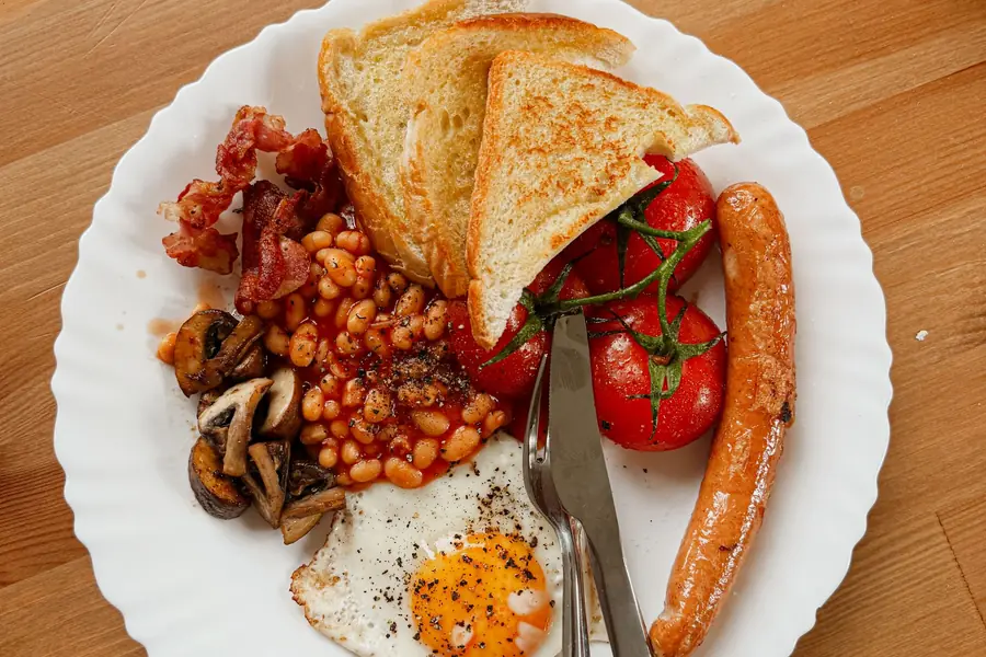 A cooked breakfast of eggs, bacon, sausage, beans, tomatoes and toast on a wooden table