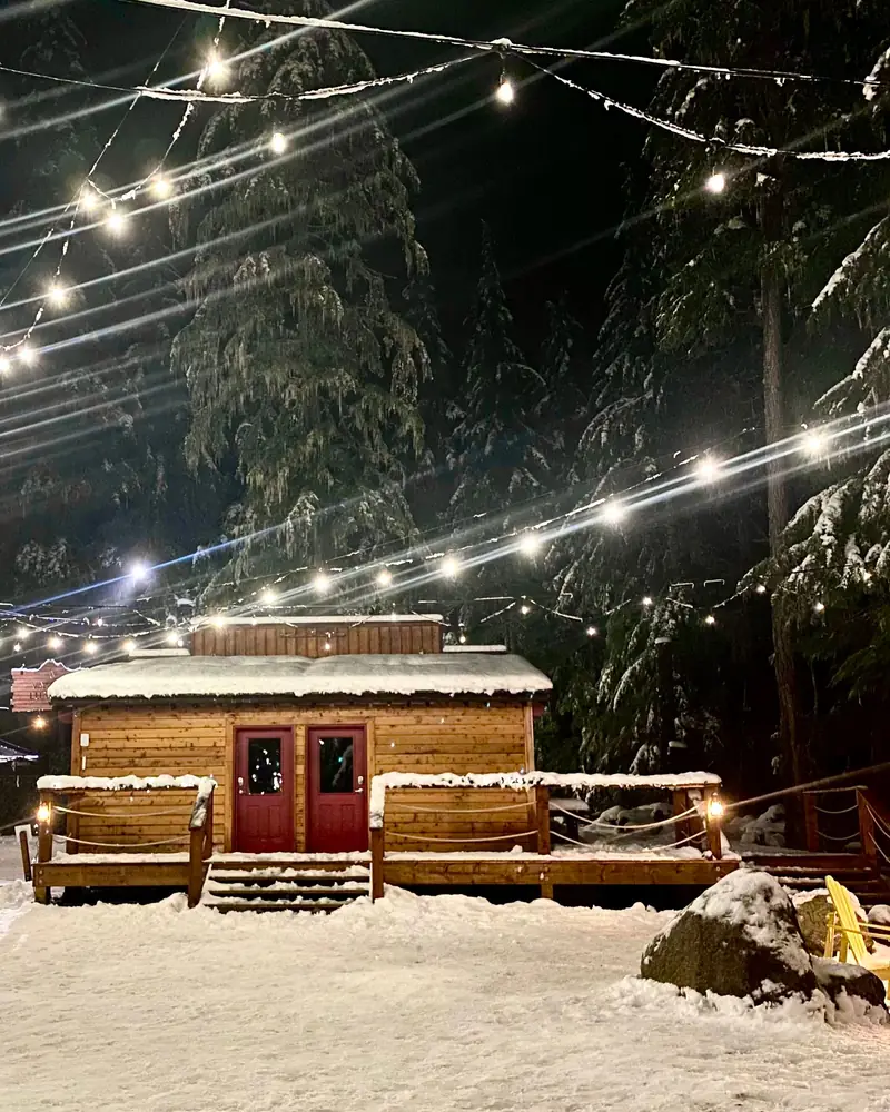 A timber cabin in the snow under strings of festoon lights at dusk