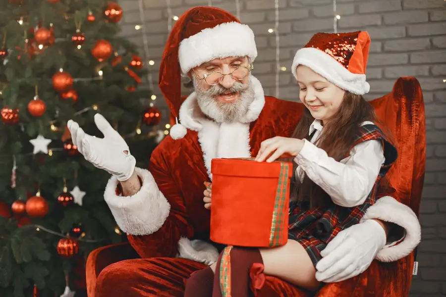 Santa helping a girl open a red present beside a Christmas tree
