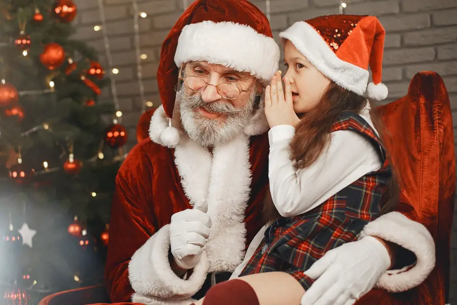 A girl in a Santa hat whispering her Christmas wish in Santa’s ear