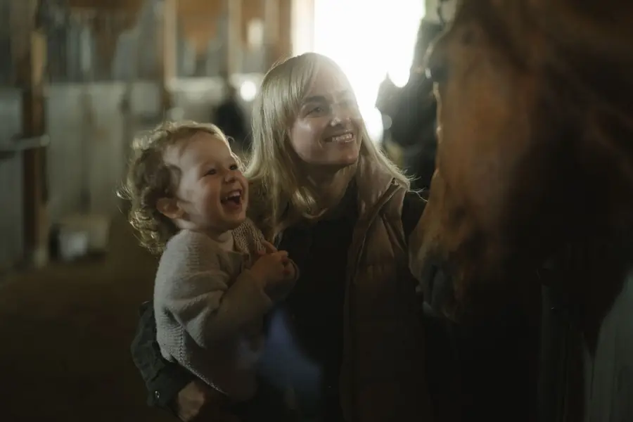 A woman holding a laughing toddler next to a horse in the stable