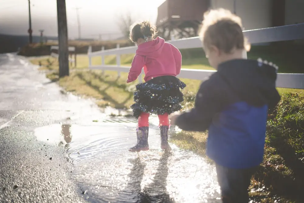 Two small children in wellies splashing in a puddle on the farm track